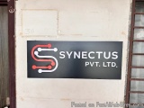 The image is a photograph of a black signboard with the logo of Synectus Pvt. Ltd. The signboard is rectangular in shape and...