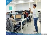 The image shows a group of people working in an office. There are six people in the image, all sitting at desks with computer...