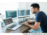 The image shows a young man sitting at a desk in front of three computer monitors and a laptop. He is wearing a blue shirt an...