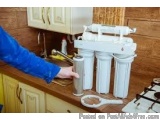 The image shows a person's hand holding a water filter in a kitchen. The filter is white and appears to be made of plastic an...