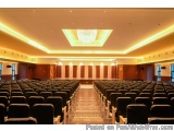 The image shows a large auditorium with rows of black chairs facing the front of the room. The room has a high ceiling with a...