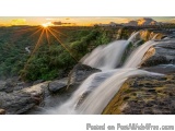 The image is a photograph of a beautiful waterfall at sunset. The waterfall is located in a mountainous area with trees and g...