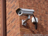 The image shows a security camera mounted on a brick wall. The camera is rectangular in shape and has a round lens on top. It...
