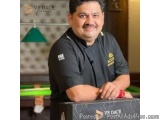 The image is a portrait of a man sitting at a desk in a restaurant. He is wearing a black chef's uniform with a gold emblem o...