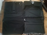 The image shows a black garment bag lying on a wooden floor. The bag is open, revealing the interior of the bag. It has multi...