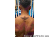 The image shows the back of a man with a tattoo on his upper back. The tattoo is of an anchor with wings and a cross on it. T...