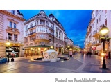The image is a photograph of a busy street in a European city at night. The street is lined with tall buildings on both sides...