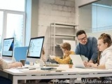The image shows a group of four people working together in an office. They are sitting around a desk with multiple computer m...