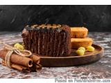 The image shows a slice of chocolate cake on a wooden plate. The cake is dark brown in color and has a thick layer of chocola...