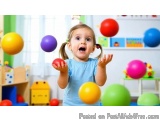 The image shows a young girl, probably around 2-3 years old, playing with colorful balls in a playroom. She is wearing a blue...