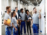 The image shows a group of six people in a factory setting, standing in front of a whiteboard and looking at it intently. The...