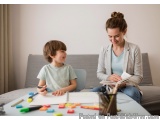 The image shows a mother and son sitting at a table in a living room. The mother is sitting on a couch and the son is sitting...