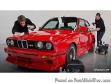 The image shows two men working on a red BMW car in a garage. The car is a BMW 3 Series E36 M3, which is a two-door coupe wit...