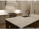 This image shows a modern kitchen with a large island in the center. The island has a white marble countertop with a sink and...