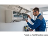 In this image, we can see a man working on an air conditioning unit mounted on the wall. He is wearing a blue uniform and gla...