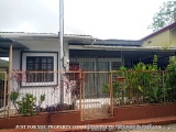 The image shows a small white house with a sloping roof and a metal fence in front of it. The house has a small window on the...