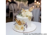 The image is of a three-tiered white cake on a white cake stand. The cake is decorated with white roses and gold leaves on to...