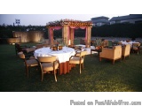 The image shows an outdoor dining area set up for a formal event. The area is set up on a grassy lawn with a stone wall in th...