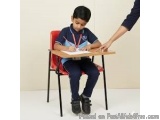 The image shows a young boy sitting at a desk with a red chair. He is wearing a blue polo shirt and black pants. He has a med...