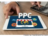 The image shows a person's hand holding a tablet with the word "PPC" on it. The tablet is placed on a wooden desk with a calc...
