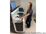 The image shows a young woman standing in front of a large printing machine in a laboratory. She is wearing a blue t-shirt an...