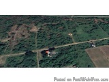 The image is an aerial view of a rural area. The area is covered in green trees and shrubs, and there are several roads windi...