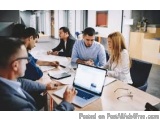 The image shows a group of six people sitting around a conference table in a meeting room. They are engaged in a discussion a...