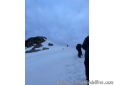 The image shows a group of people hiking up a snowy mountain. The sky is cloudy and the mountain appears to be steep and rock...