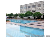 The image shows a large swimming pool in front of a tall, white building. The building appears to be a hotel or apartment com...