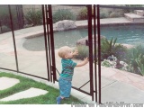The image shows a young child, probably around 2-3 years old, standing in front of a black metal gate. The child is wearing a...