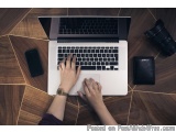 The image shows a person's hands typing on a laptop keyboard on a wooden desk. The laptop is open and the screen is turned on...