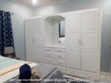 The image shows a bedroom with a large white wardrobe in the center. The wardrobe has multiple doors and drawers, and a mirro...