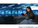 The image shows a woman and a man in a server room with multiple computer monitors. The woman is standing in front of the mon...