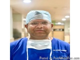 The image is a portrait of a man wearing a blue surgical gown and a surgical cap. He is also wearing a face mask and glasses....