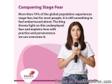 The image shows a young woman holding a microphone and speaking. She is wearing a white t-shirt and has long brown hair. The...
