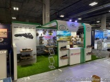The image shows a trade show booth with a green and white color scheme. The booth has a large banner on the left side with an...