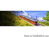 The image shows a red train traveling on a railway track in a mountainous area. The train is moving towards the right side of...
