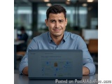 The image shows a young man sitting at a desk with a laptop in front of him. He is wearing a blue collared shirt and has a se...