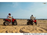 The image shows two people riding red ATVs on a sandy beach. The person on the left is wearing a white shirt and a red helmet...