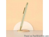 The image shows a gold-colored pen resting on a wooden stand. The pen has a sleek and modern design with a pointed tip and a...