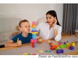 The image shows a mother and son playing with colorful building blocks on a wooden table. The mother is sitting on the right...