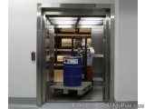 The image shows an open elevator in a warehouse or industrial setting. The elevator is made of stainless steel and has a glas...