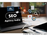 The image shows a laptop with the words "SEO Agency India" written on the screen. The laptop is placed on a desk with papers...
