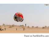 The image shows a group of people parasailing in a desert-like area. The sky is clear and blue, and the ground is dry and bar...