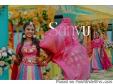 The image shows a young girl wearing a traditional Indian outfit. She is standing in front of a colorful backdrop with flower...