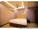 The image shows a modern bedroom with a large bed in the center. The bed has a wooden frame and a beige tufted headboard. The...