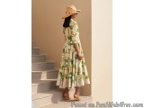 The image shows a woman standing on a set of stairs. She is wearing a long, flowy dress with a green and white tropical print...