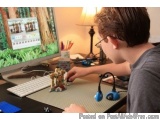 The image shows a young boy sitting at a desk in front of a computer monitor. He is wearing a gray t-shirt and glasses and ap...