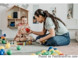 The image shows a mother and daughter playing with colorful wooden blocks in a playroom. The mother is sitting on the floor a...