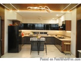 The image shows a modern kitchen with a large island in the center. The kitchen has a black refrigerator on the left side and...
